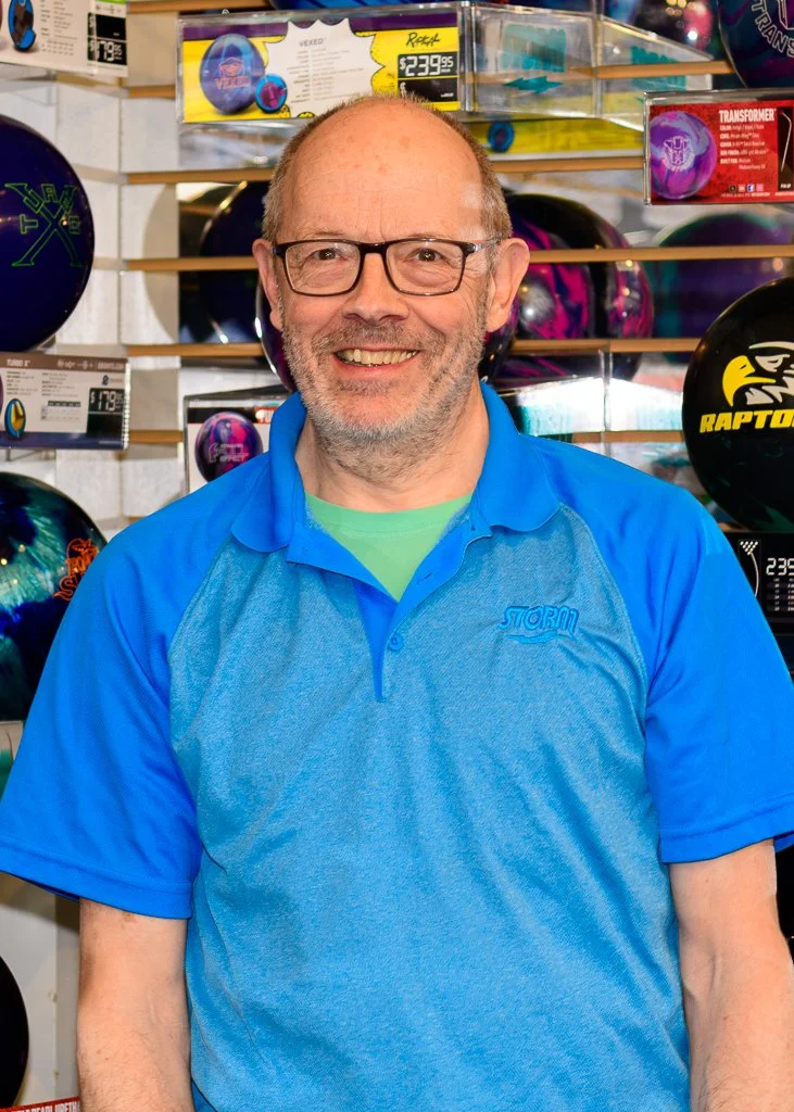 A smiling man wearing glasses and a blue collared shirt stands in a bowling alley with bowling balls on shelves behind him.