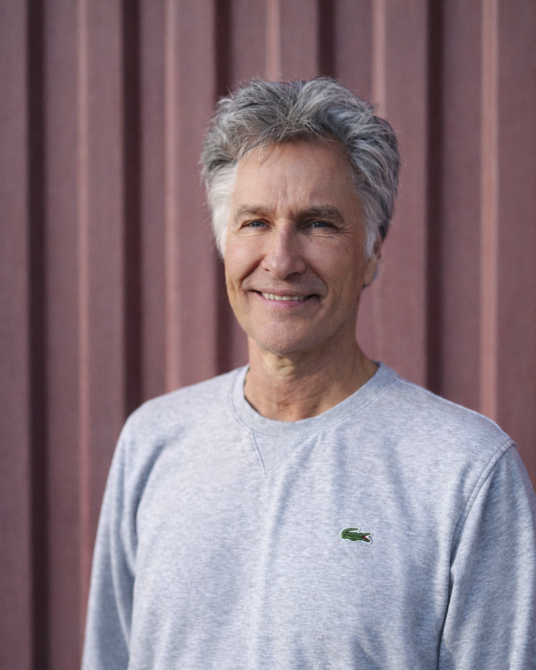 A middle-aged man with gray hair smiling, wearing a gray Lacoste sweatshirt, standing in front of a wooden fence.