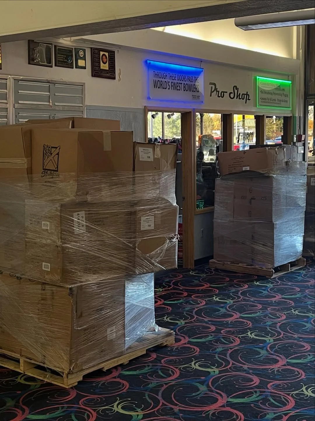 Store interior with cardboard boxes on pallets wrapped in plastic, neon signs, and a patterned carpet.