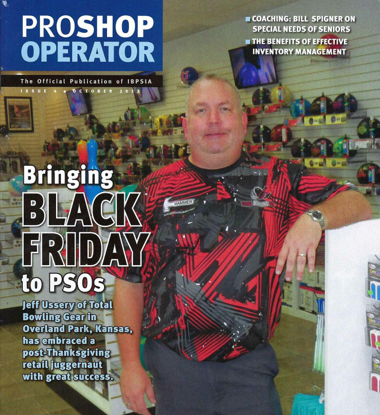 Man in red and black shurt standing inside a store with shelves of bowling balls, next to a white counter.