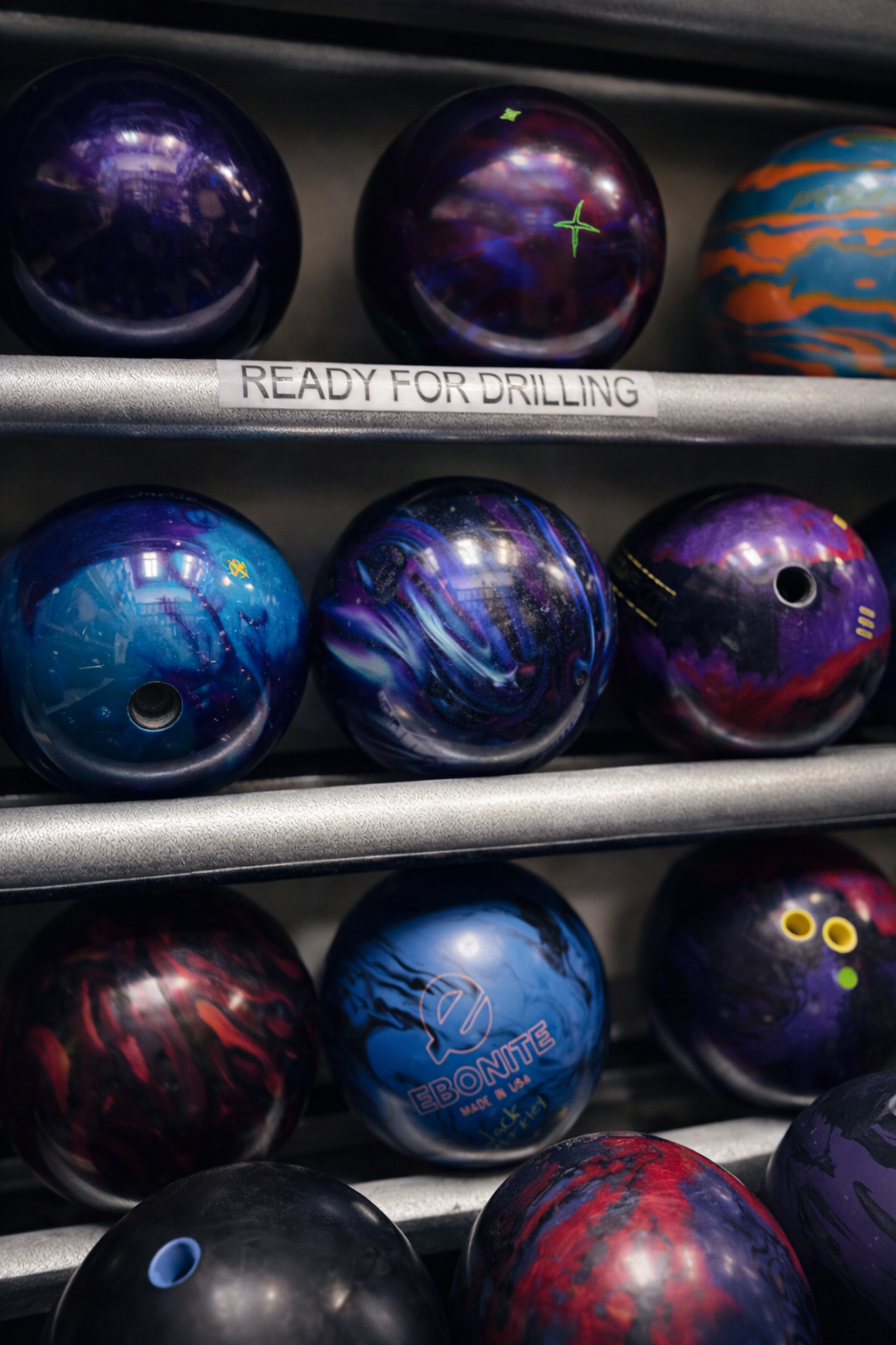 Bowling balls of various colors and patterns on a metal rack, with a label reading 'READY FOR DRILLING'.