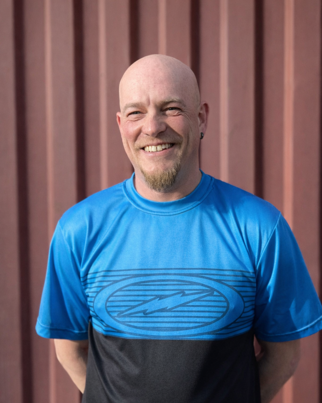 A smiling man with a goatee and earring stands in front of a wooden background, wearing a blue and black athletic shirt.