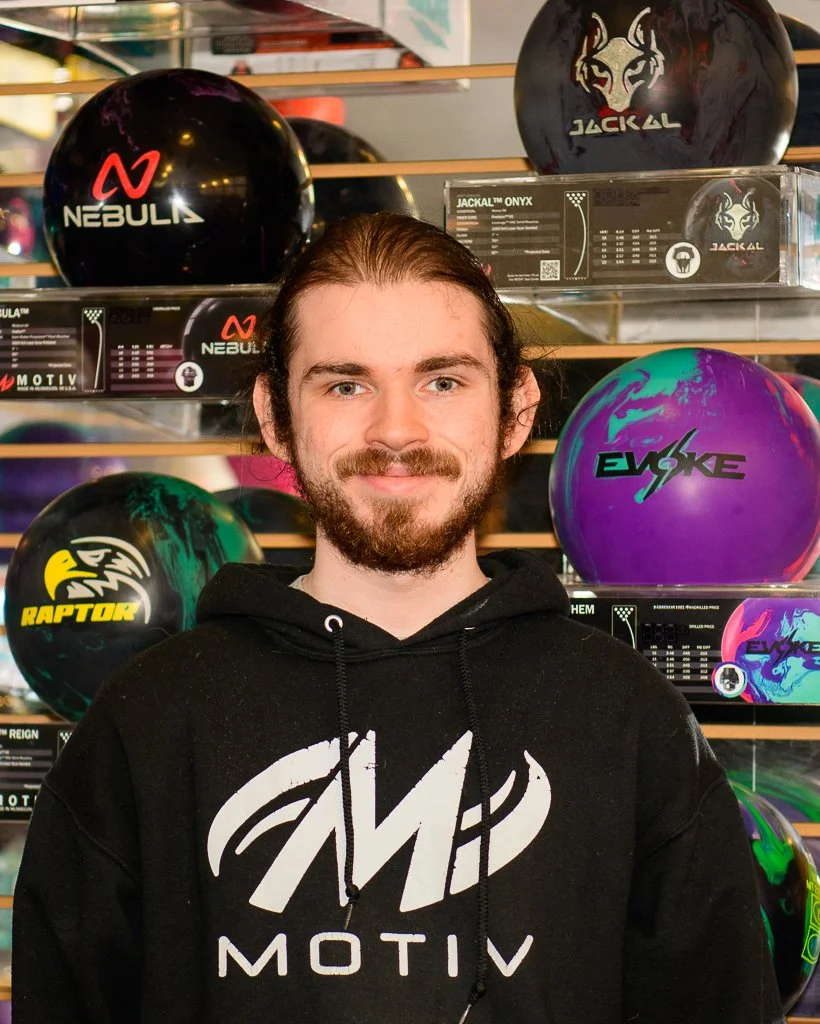 A young man with long brown hair, a beard, and a mustache, smiling, wearing a black hoodie with a white MOTIV logo, standing in front of a display of colorful bowling balls in a store.