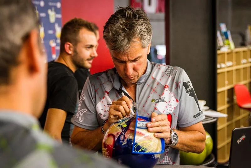 A man with gray hair signing a bowling ball at a store or event. Two other men are in the background, and shelves with bowls and red chairs are visible.