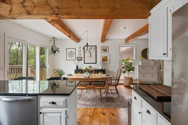 Open-concept kitchen and dining area with white walls, wooden ceiling beams, and large windows showing a view of greenery outside. The dining table is wooden with four chairs, and there is a black chandelier overhead. The space is decorated with framed artwork, plants, and a small area rug under the table.