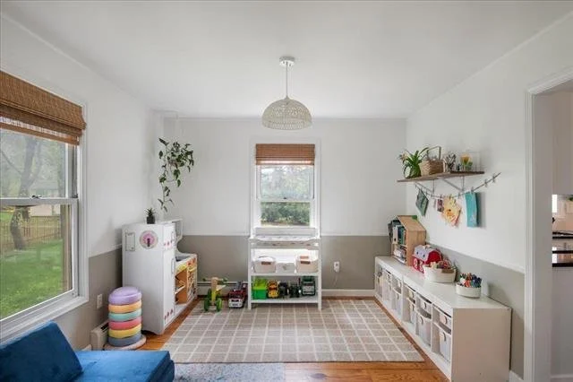 Bright children’s playroom with white walls, windows with brown blinds, and a beige checkered rug. Contains a blue couch, colorful stacked cushions, white storage units with toys, and wall shelves with books and decor. A pendant light hangs from the ceiling.