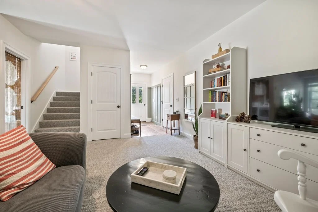 Living room with a gray couch, a black coffee table with a tray, a large flat-screen TV, white built-in shelves, and a staircase with carpeted steps.