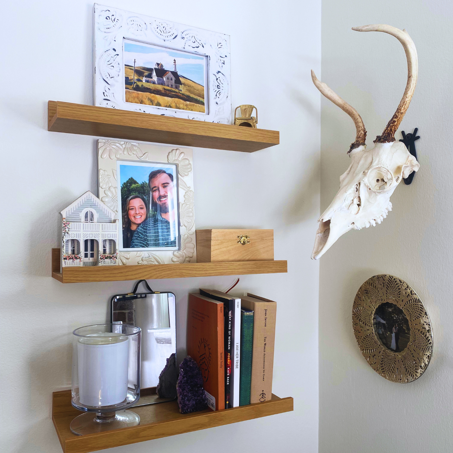 Wall-mounted wooden shelves holding framed photos, books, a small house model, a gold chair figurine, a skull of an animal with antlers, candles, purple crystals, and decorative plates.