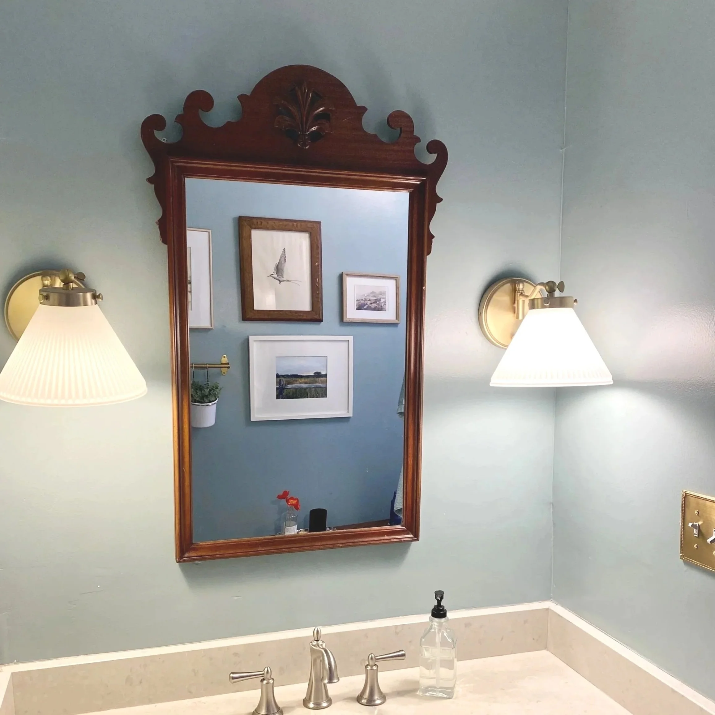 Bathroom vanity with a mirror, two wall-mounted lamps, framed artwork, a plant, and soap dispenser on the countertop.
