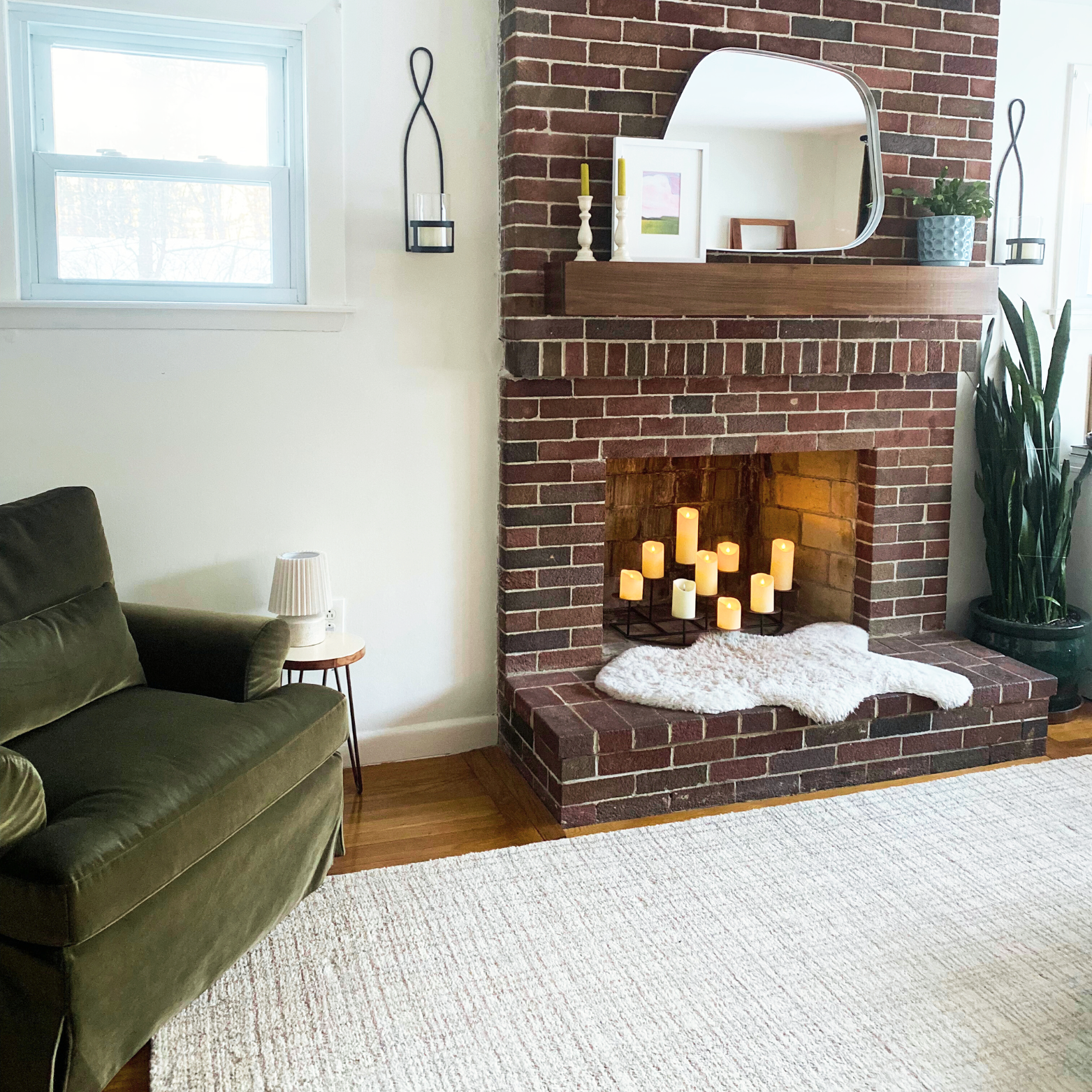 Living room with brick fireplace decorated with candles, framed artwork, and plants, alongside a green sofa and a side table with a lamp, and a large window letting in natural light.