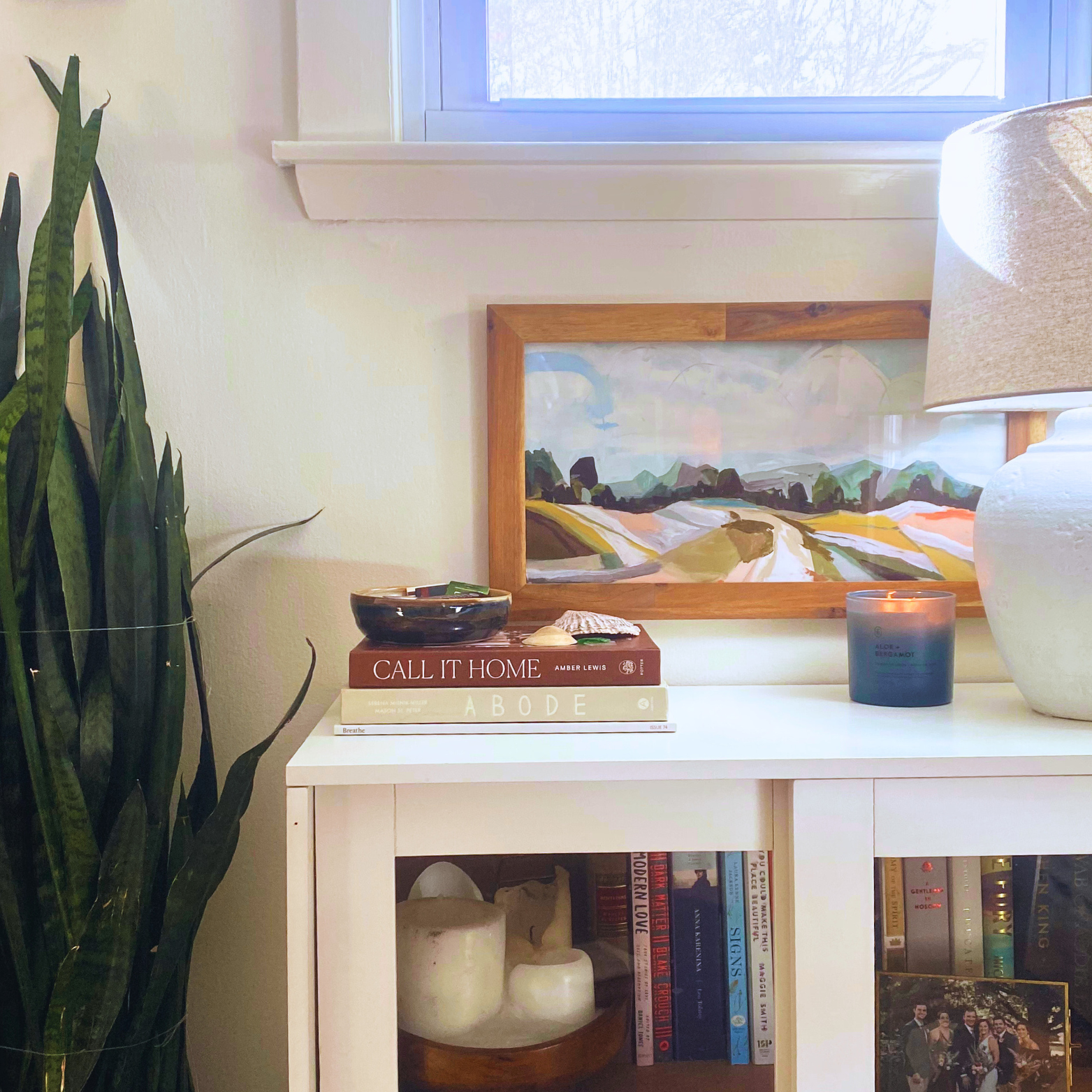 A white bookshelf with books and decorative items, a framed landscape painting, a candle, and a large white vase on top, with a window and a houseplant nearby.