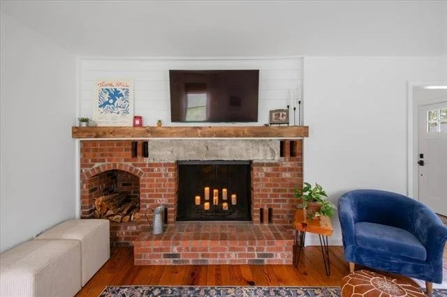 Living room fireplace with brick and stone, a wall-mounted TV above, and a wooden mantel with decor, a blue armchair, a small table with a green plant, and a white door to the right.