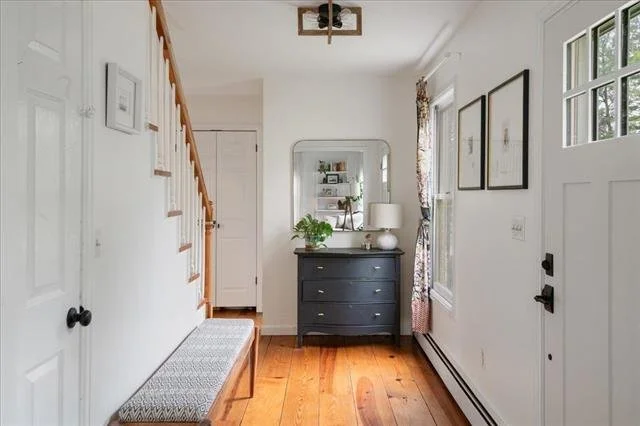 View of an entryway with a staircase on the left, a dark dresser with a mirror and lamp in the center, and a door and window with floral curtains on the right. The floor is hardwood and walls are painted white.