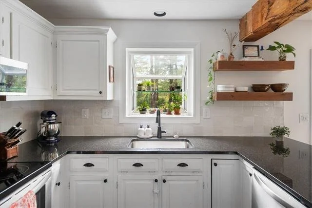 Bright kitchen with white cabinets, black countertops, a window with greenery outside, and wooden open shelves with bowls and plants.