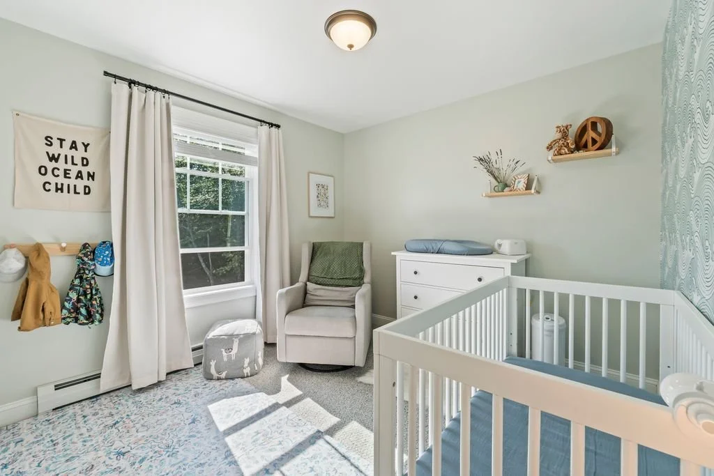 A nursery room with a white crib, a white dresser, a gray armchair with a green blanket, a window with white curtains, and wall decorations including a sign that reads "Stay Wild Ocean Child" and floating shelves with decorative items.