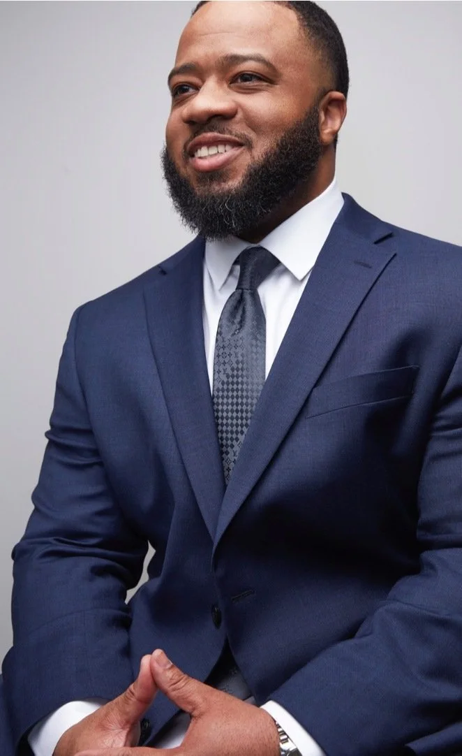 A confident black man in a navy suit, white shirt, and patterned tie, smiling and standing against a neutral background.