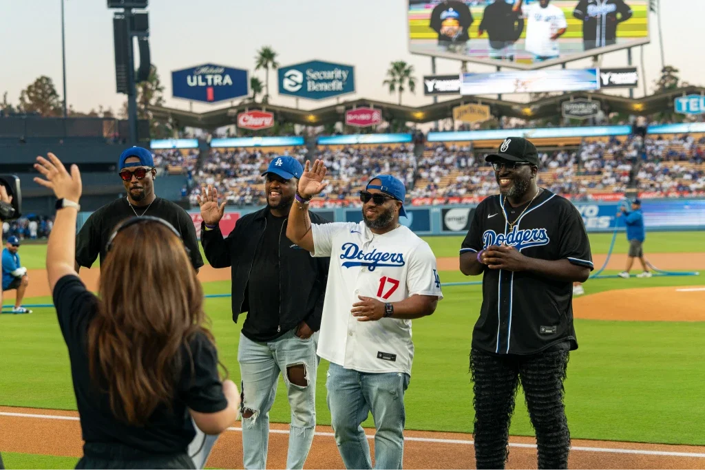 Four men on a baseball field, three are wearing Dodgers jerseys and caps, and one woman with long hair is facing them with her hand raised in greeting, likely during a sports event.