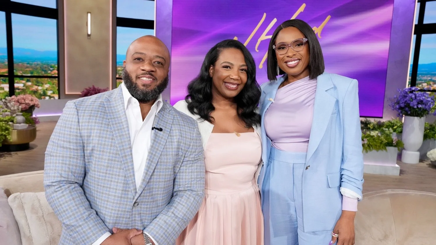 Three TV show hosts standing together in a studio with a cityscape view and purple backdrop.