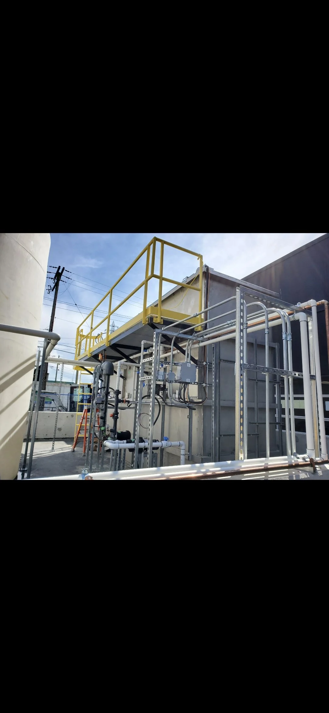 Industrial piping system with metal framework, electrical boxes, and a yellow safety railing outdoors under a blue sky.