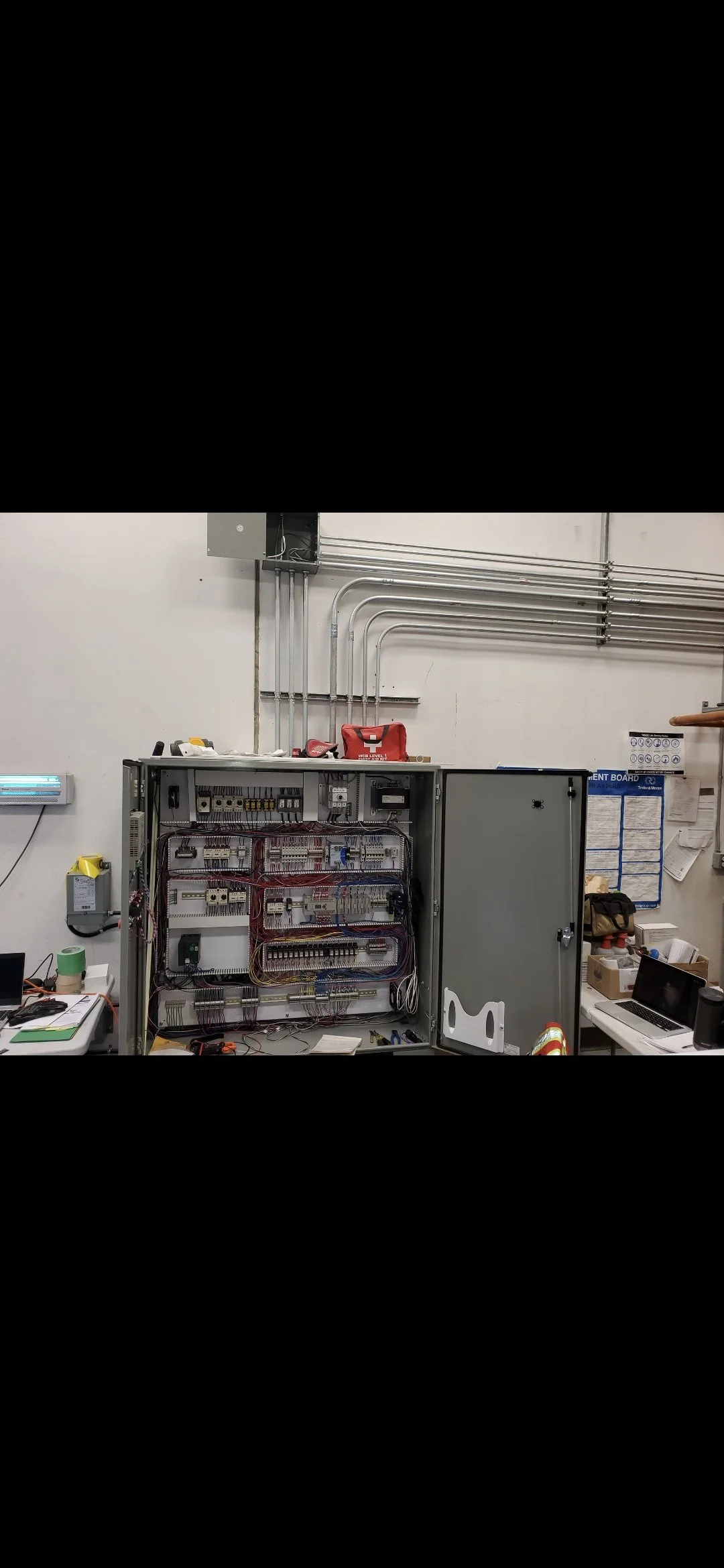 An electrical control panel with various wiring and components, situated in an industrial or utility room, with tools and equipment nearby.