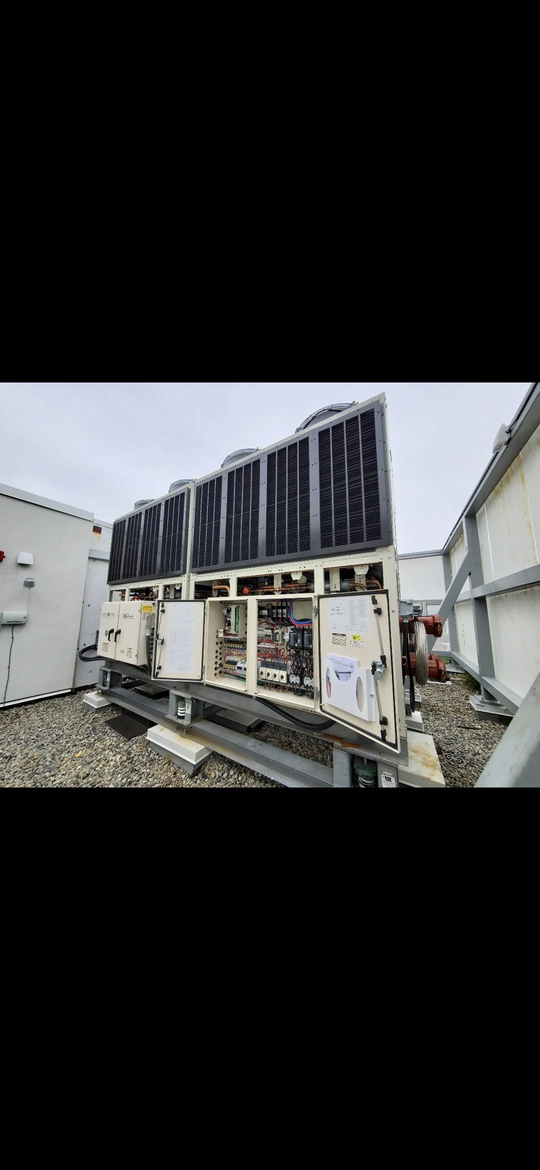 Large industrial HVAC or refrigeration system with multiple black cooling fins and open electrical panels, installed outdoors on a gravel surface under a cloudy sky.