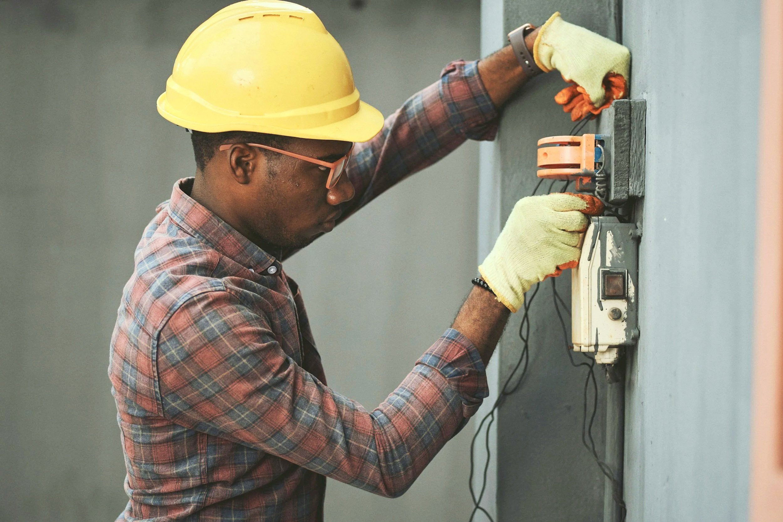 A man wearing a yellow safety helmet, glasses, gloves, and a plaid shirt is working on an electrical panel or device mounted on a wall. He is using tools and appears to be repairing or installing the electrical equipment.