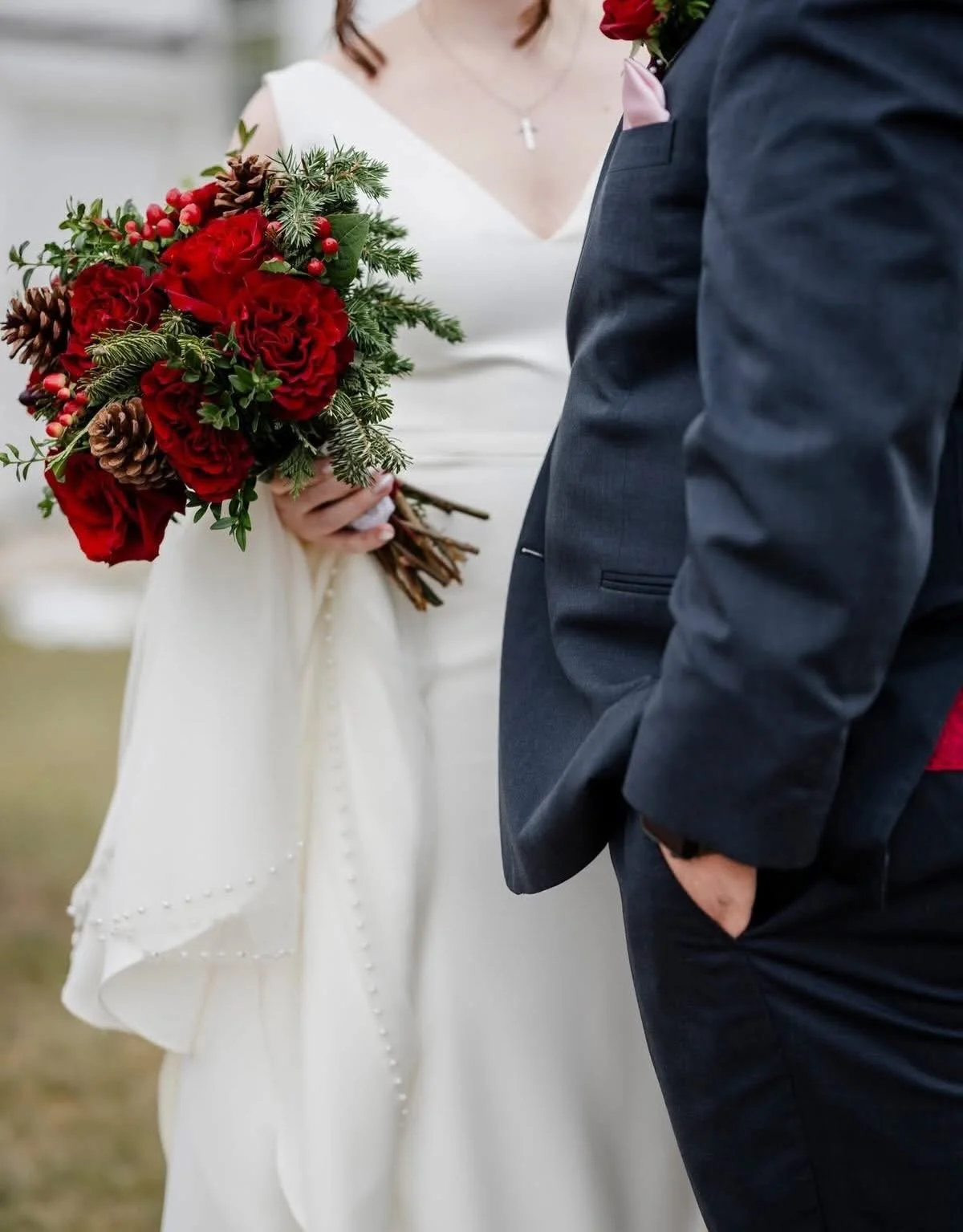 A bride holding a bouquet of red flowers, pine cones, and greenery stands next to a groom wearing a dark suit with a pink boutonniere. The bride is dressed in a white gown with pearl embellishments, and the groom has his hand in his pocket.
