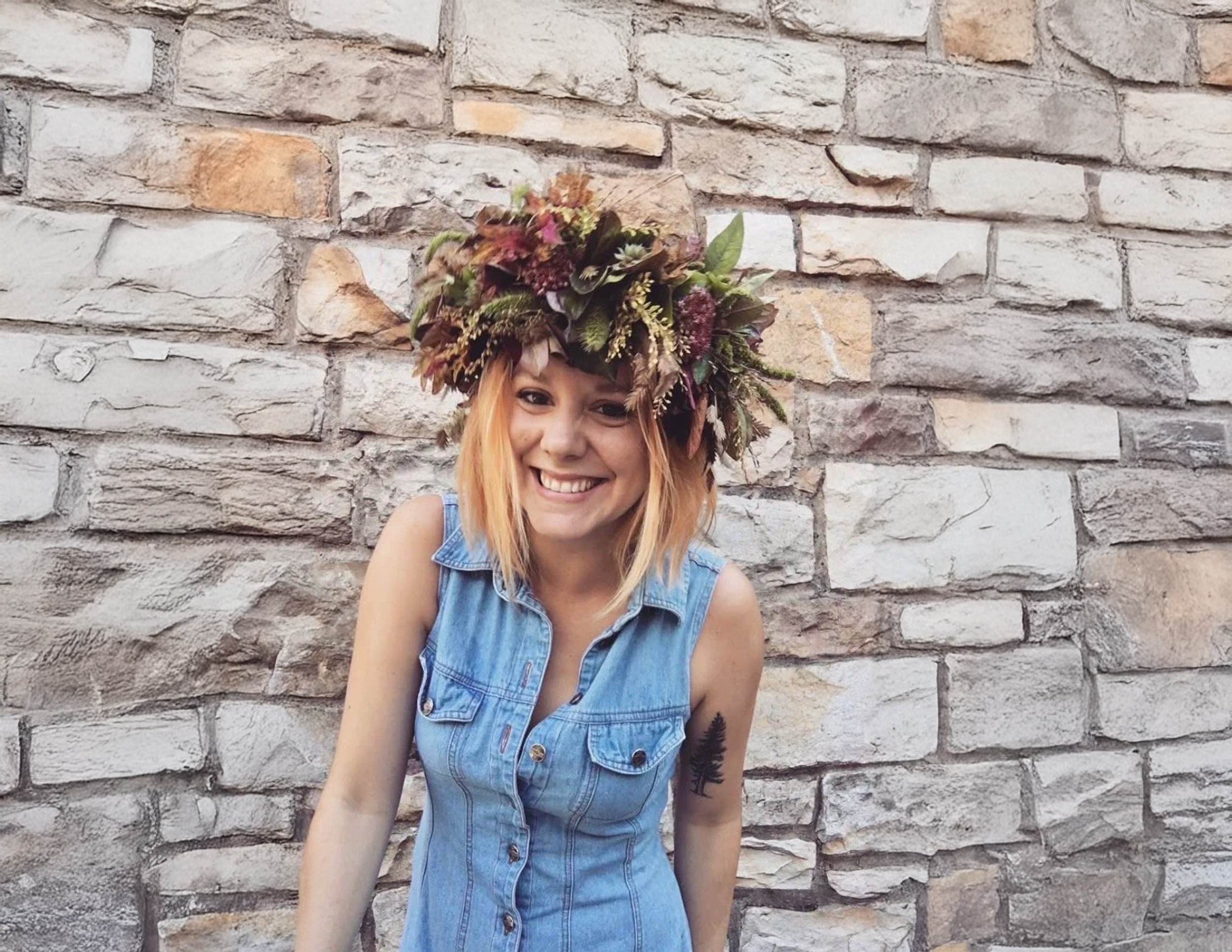 A woman with blonde hair and a floral crown smiling in front of a stone wall.