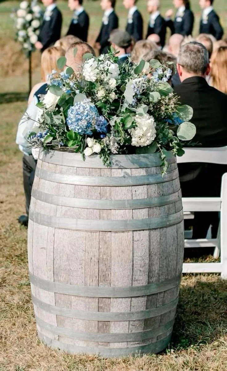 A large floral arrangement of white and blue flowers with green foliage in a wine barrel at an outdoor wedding ceremony.