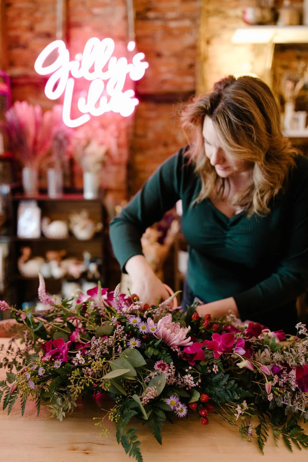 A woman arranging pink and purple flowers on a wooden table in a floral shop, with a neon sign reading 'Gilly's Love' in the background.