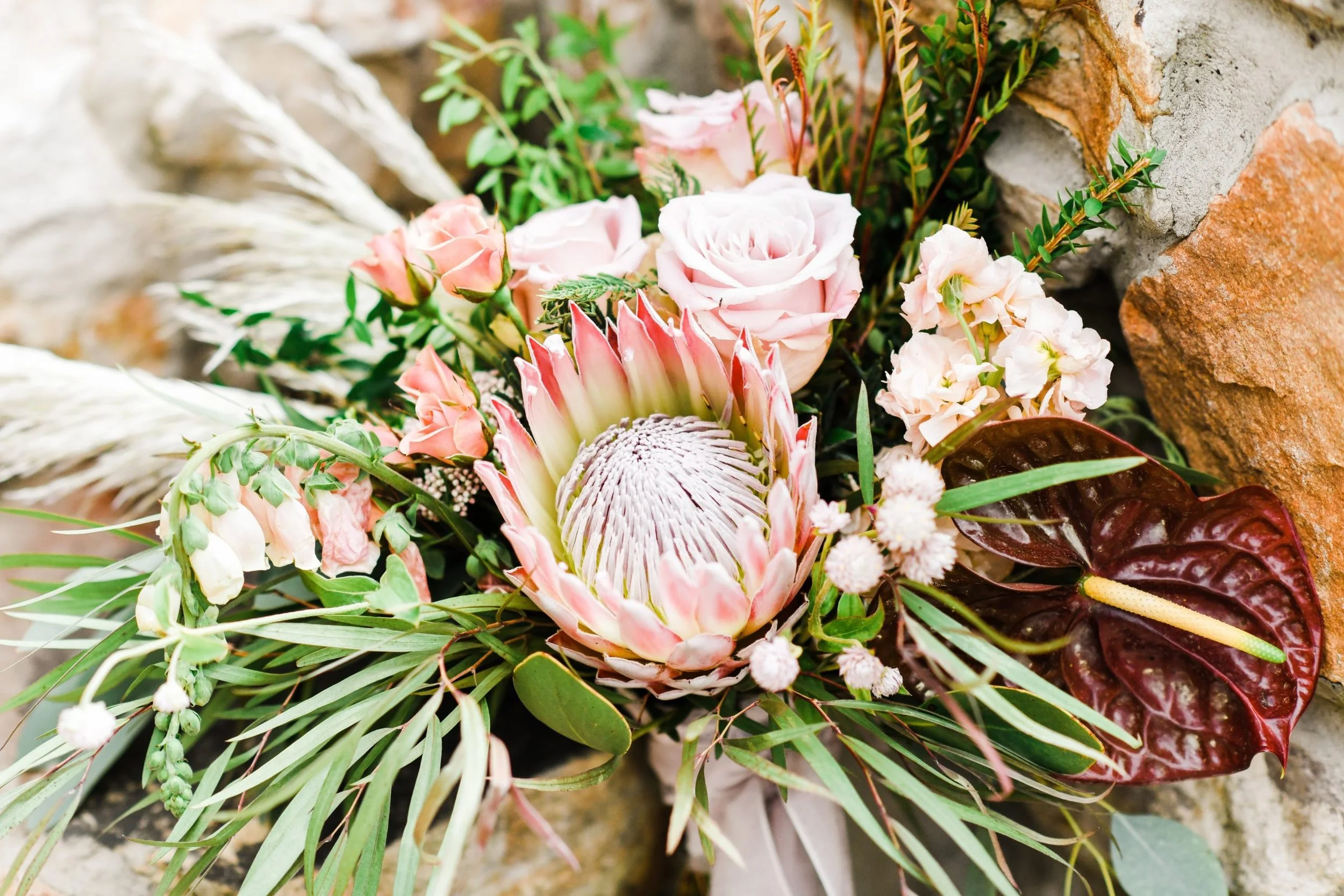 A floral arrangement featuring pink roses, a large protea, white and pink small flowers, green foliage, and a red anthurium, placed against a brick and stone background.