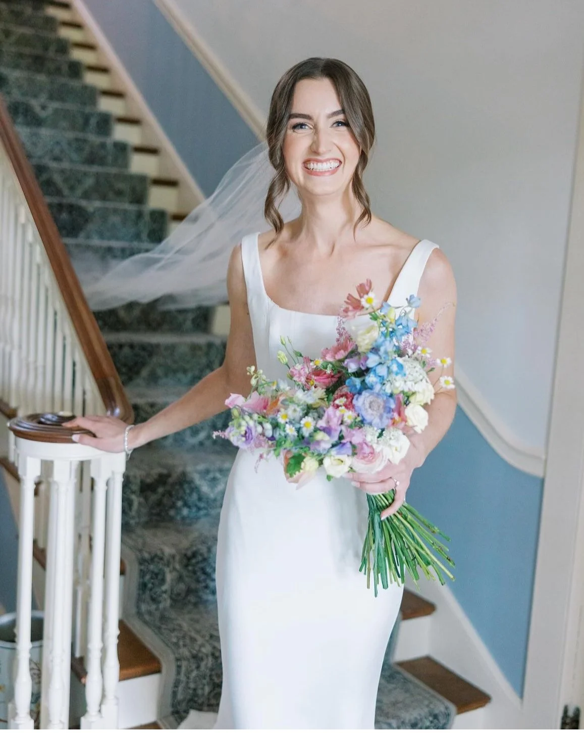 Bride in a white wedding dress holding a colorful bouquet, standing on a staircase, smiling with a veil flowing behind her.