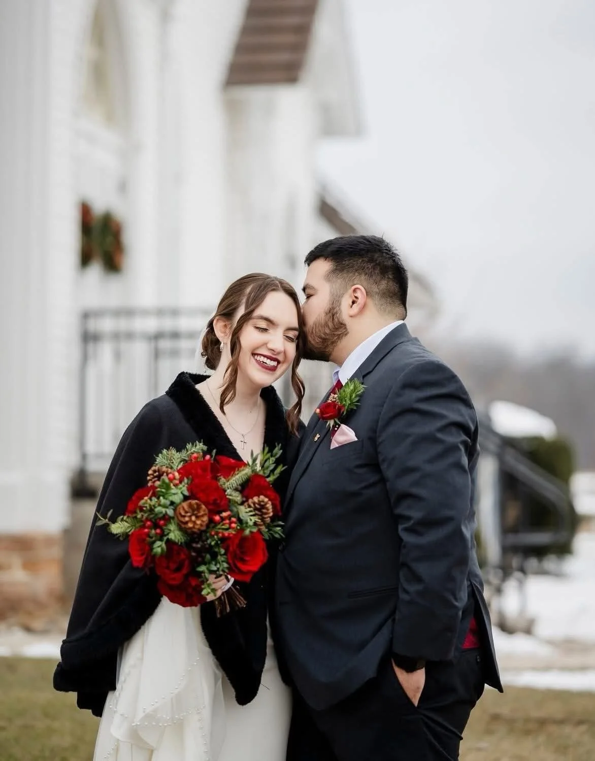 A newlywed couple sharing a kiss outside on a cloudy day, with the groom kissing the bride's forehead. The bride is holding a bouquet of red flowers and pinecones, and both are dressed in formal wedding attire. The background features a white building with black accents and snow on the ground.