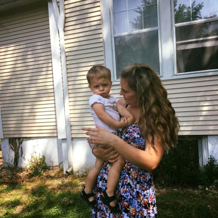 A woman holding a young girl outside a house with beige siding and windows.