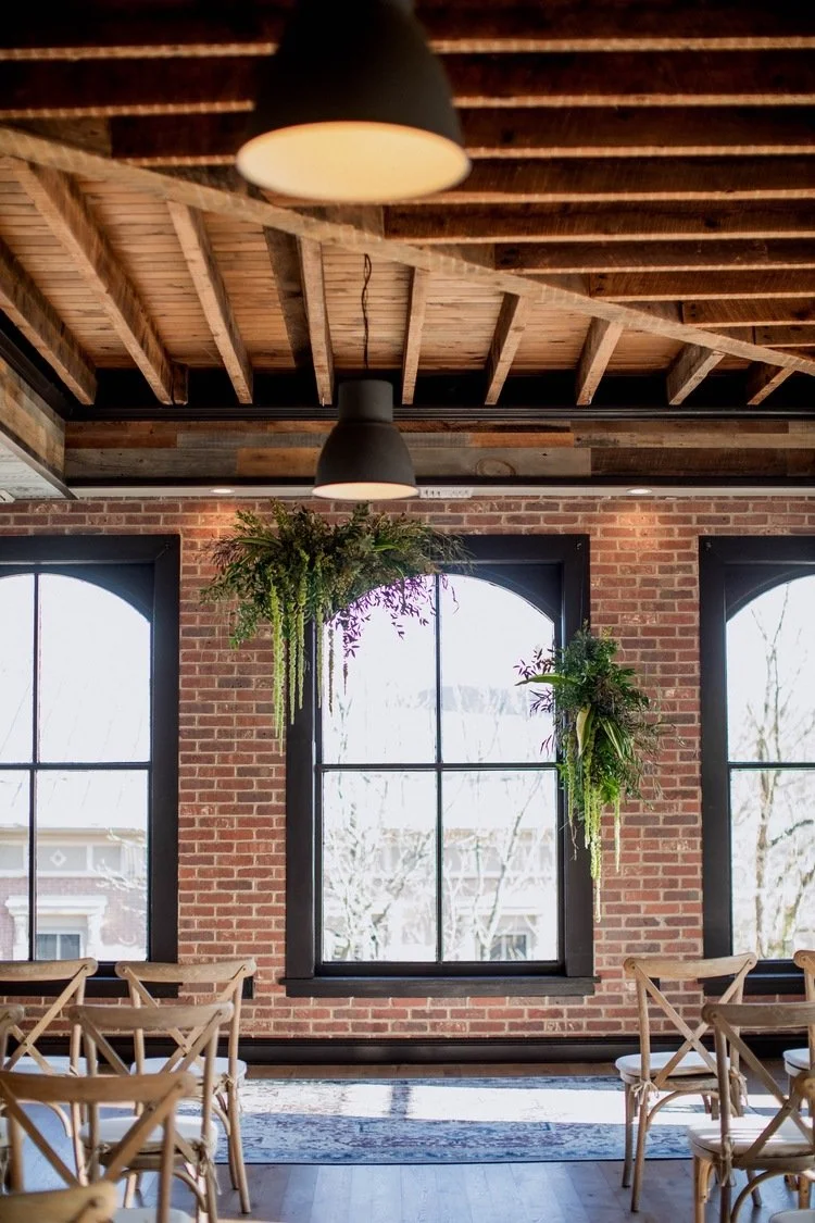 Interior of a room with exposed brick walls and large arched windows, decorated with hanging green plants, wooden chairs, and pendant lights.
