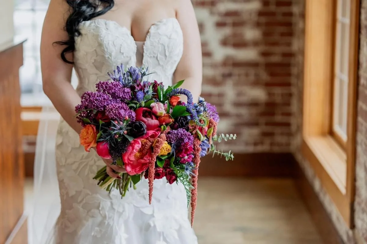 A bride in a white lace wedding gown holding a large, colorful bouquet of flowers, standing inside a room with wooden floors and brick walls.