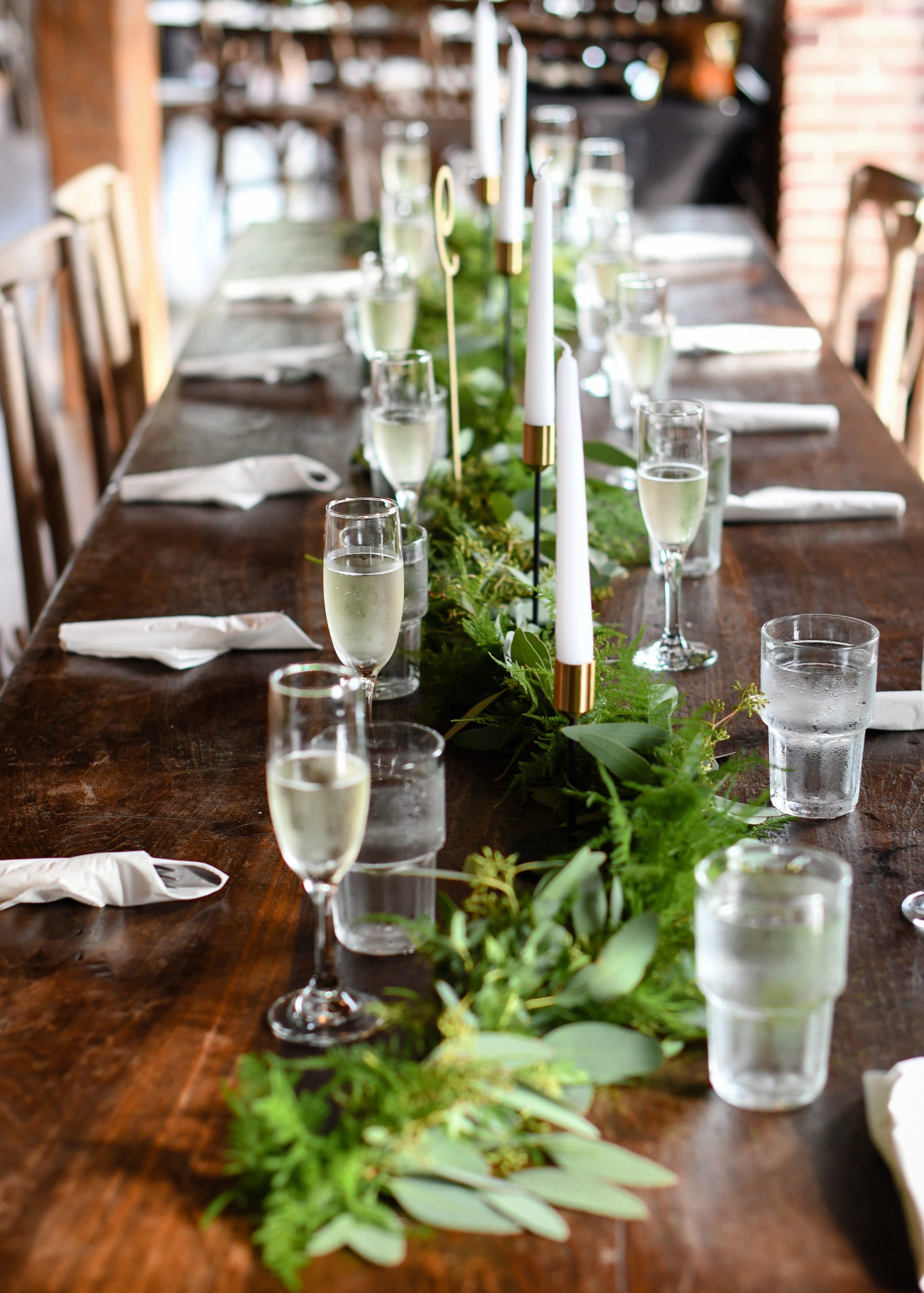 A long wooden dining table decorated with green foliage and white candles in gold and black holders, set with champagne glasses and water glasses, and white napkins at each seat.