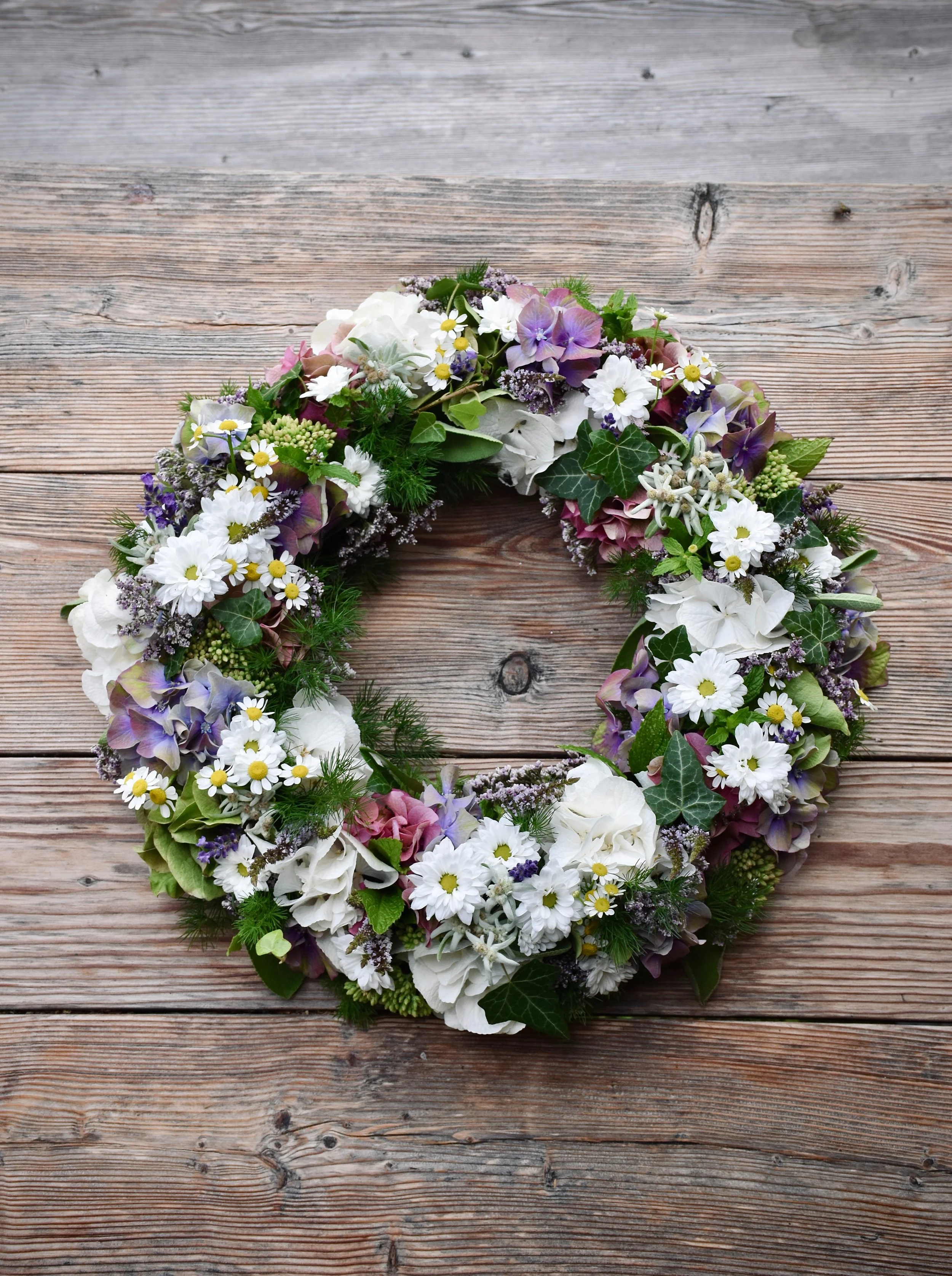 A colorful flower wreath with white, purple, and pink flowers, accented with green foliage, placed on a wooden surface.