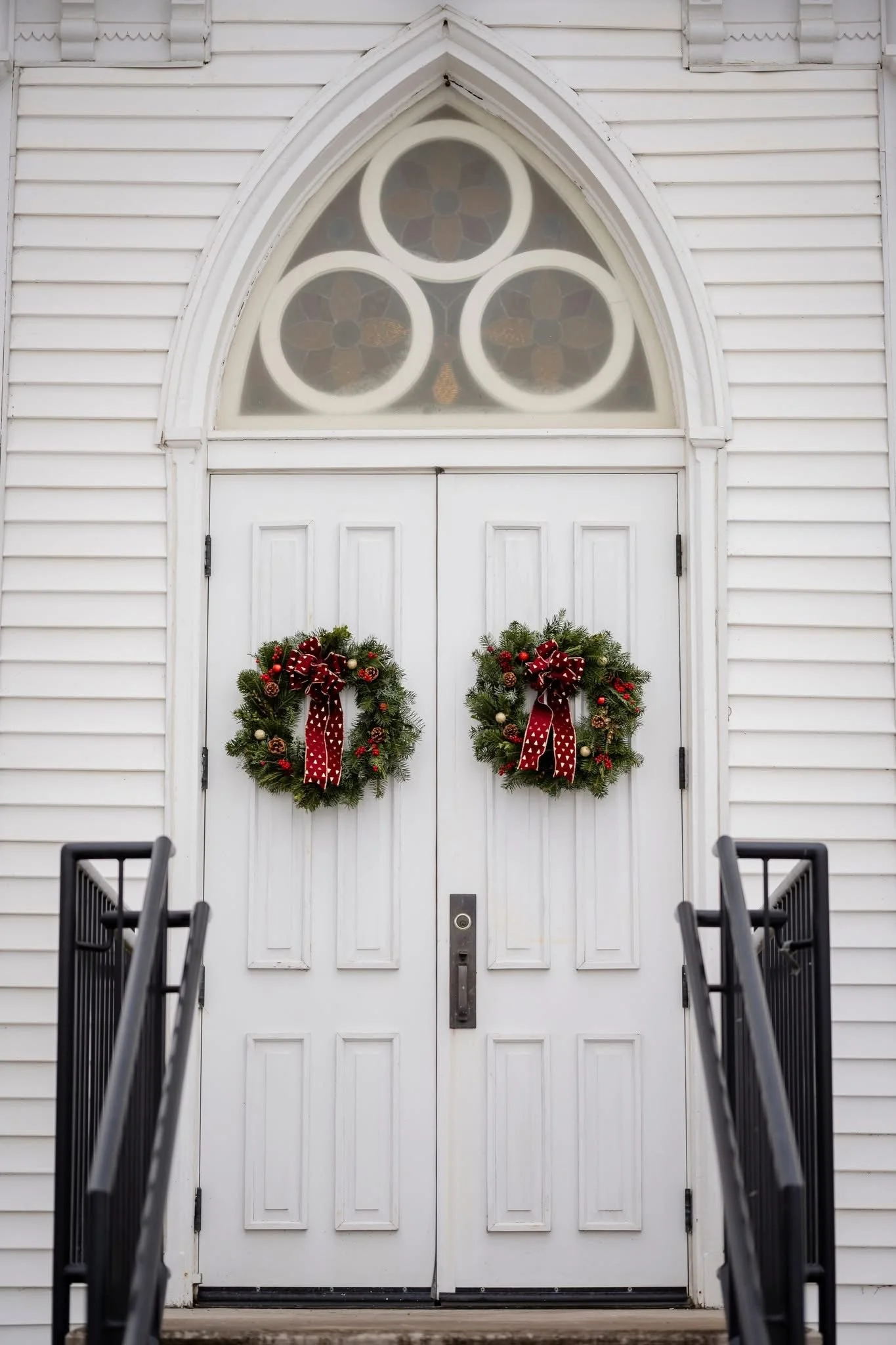 White church door decorated with two Christmas wreaths with red ribbons, wreaths on a white wooden building with a stained glass window above.