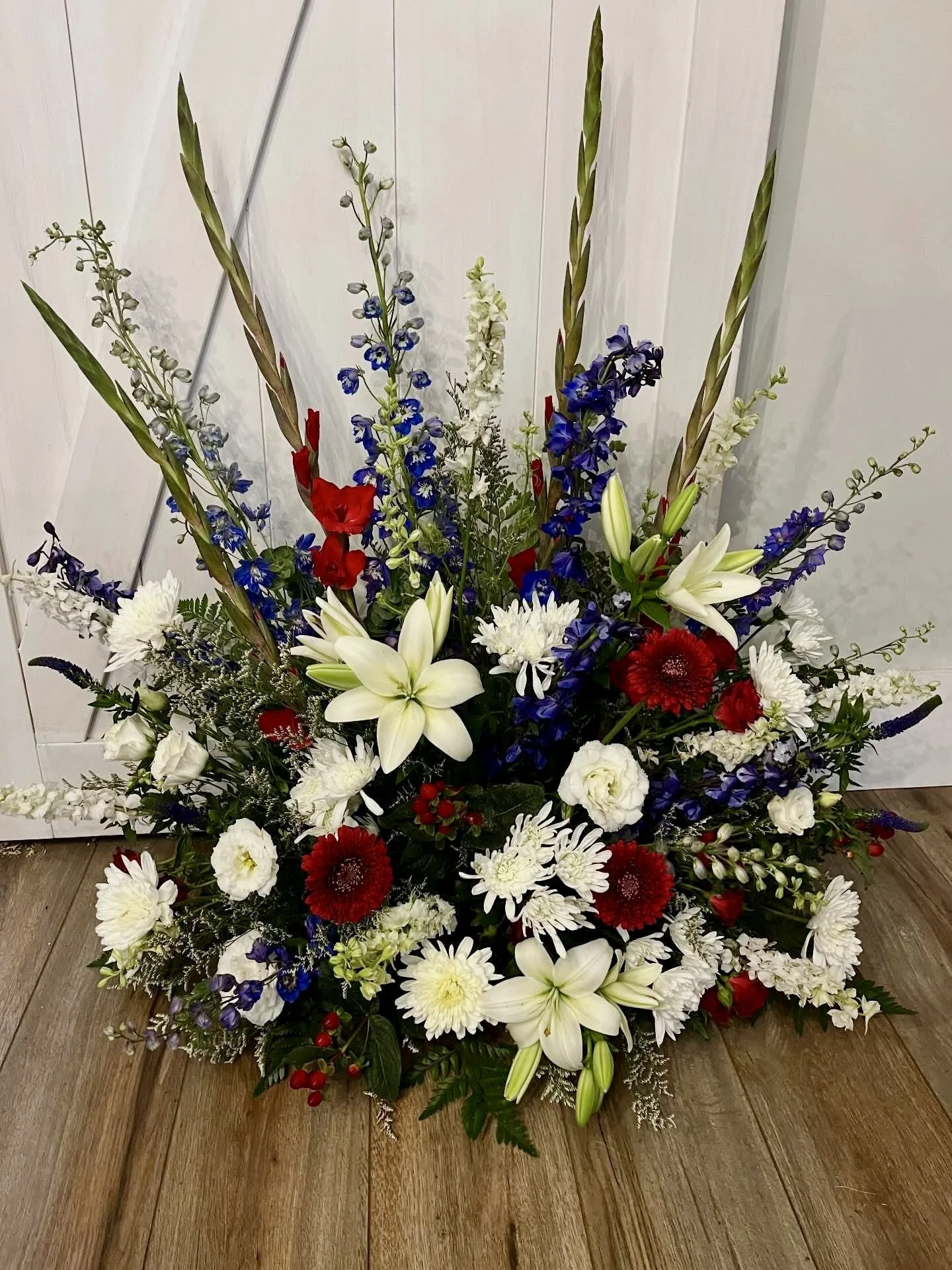 A colorful floral arrangement with white lilies, red and white daisies, blue delphiniums, and other flowers, set on a wooden floor against a white panel background.
