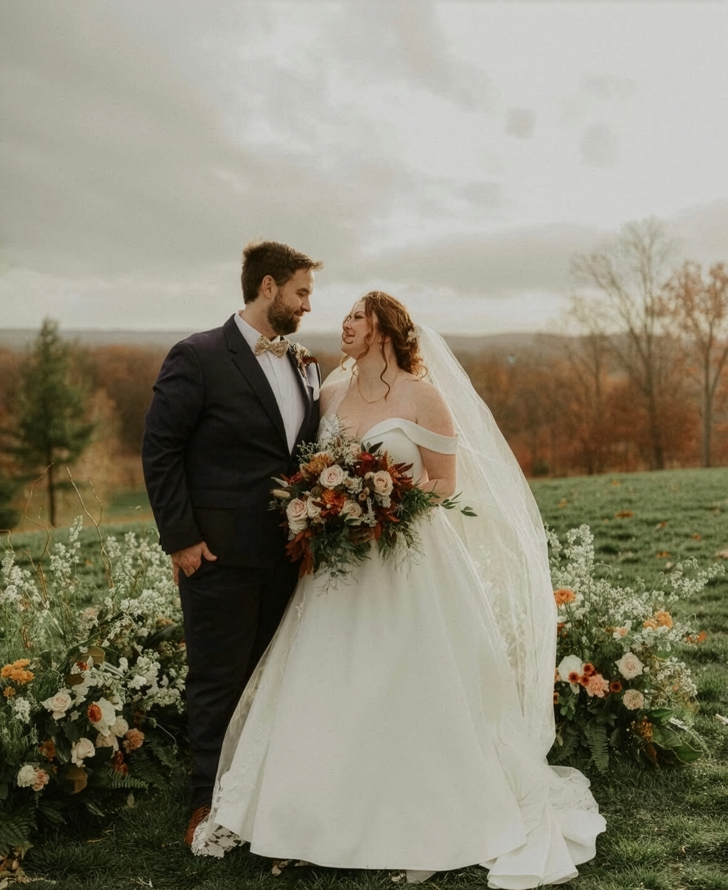 Wedding couple standing outdoors with floral arrangements, the groom in a black suit and bow tie, the bride in a white gown holding a bouquet, scenic countryside background with trees and cloudy sky.
