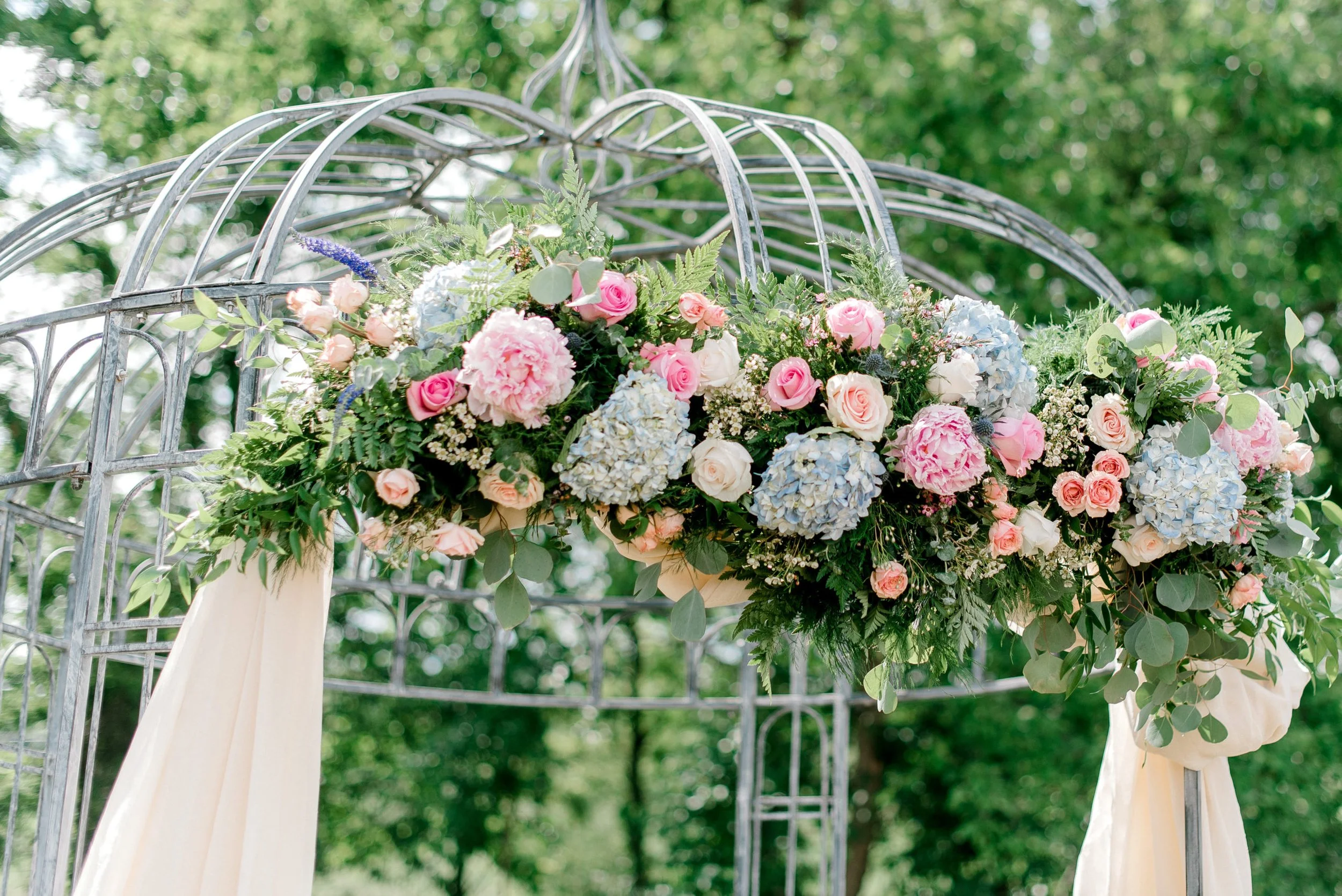 Decorative floral arrangement with pink and white roses, blue hydrangeas, and greenery on a metal archway outdoors.