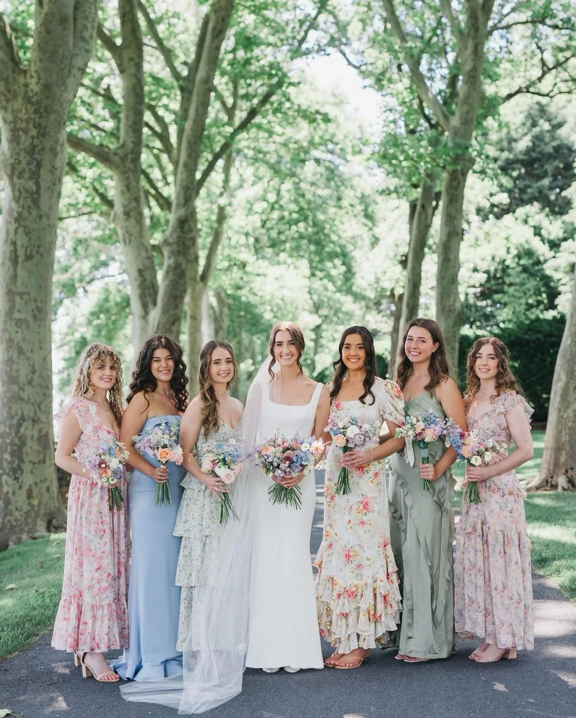 A group of seven women, including a bride in a white wedding dress, standing outdoors under large trees on a sunny day, all holding floral bouquets and smiling.