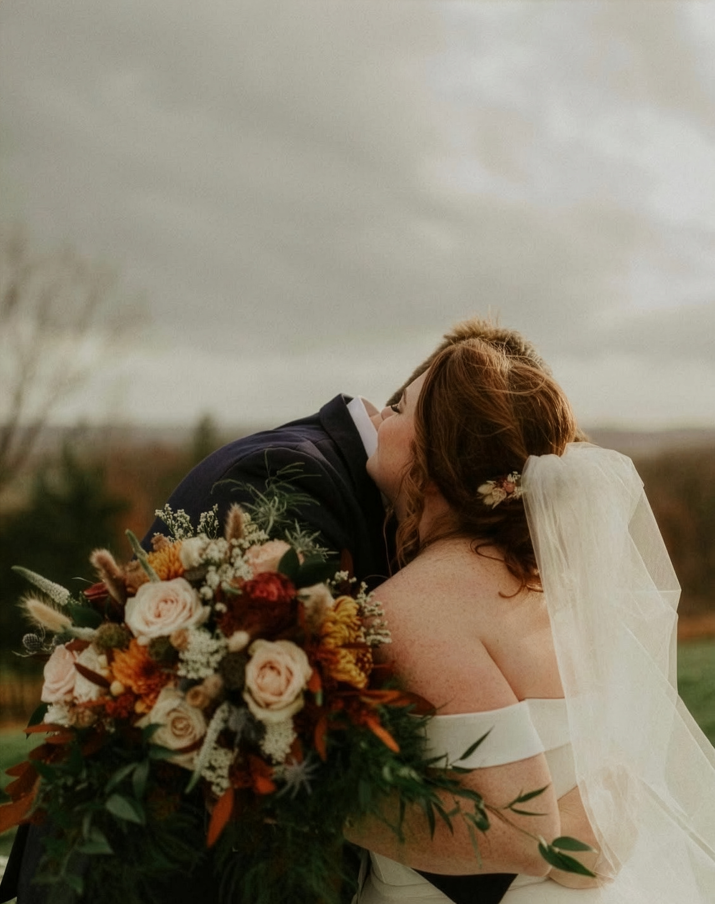 Bride and groom sharing a kiss outdoors on their wedding day, with the bride holding a large bouquet of flowers.