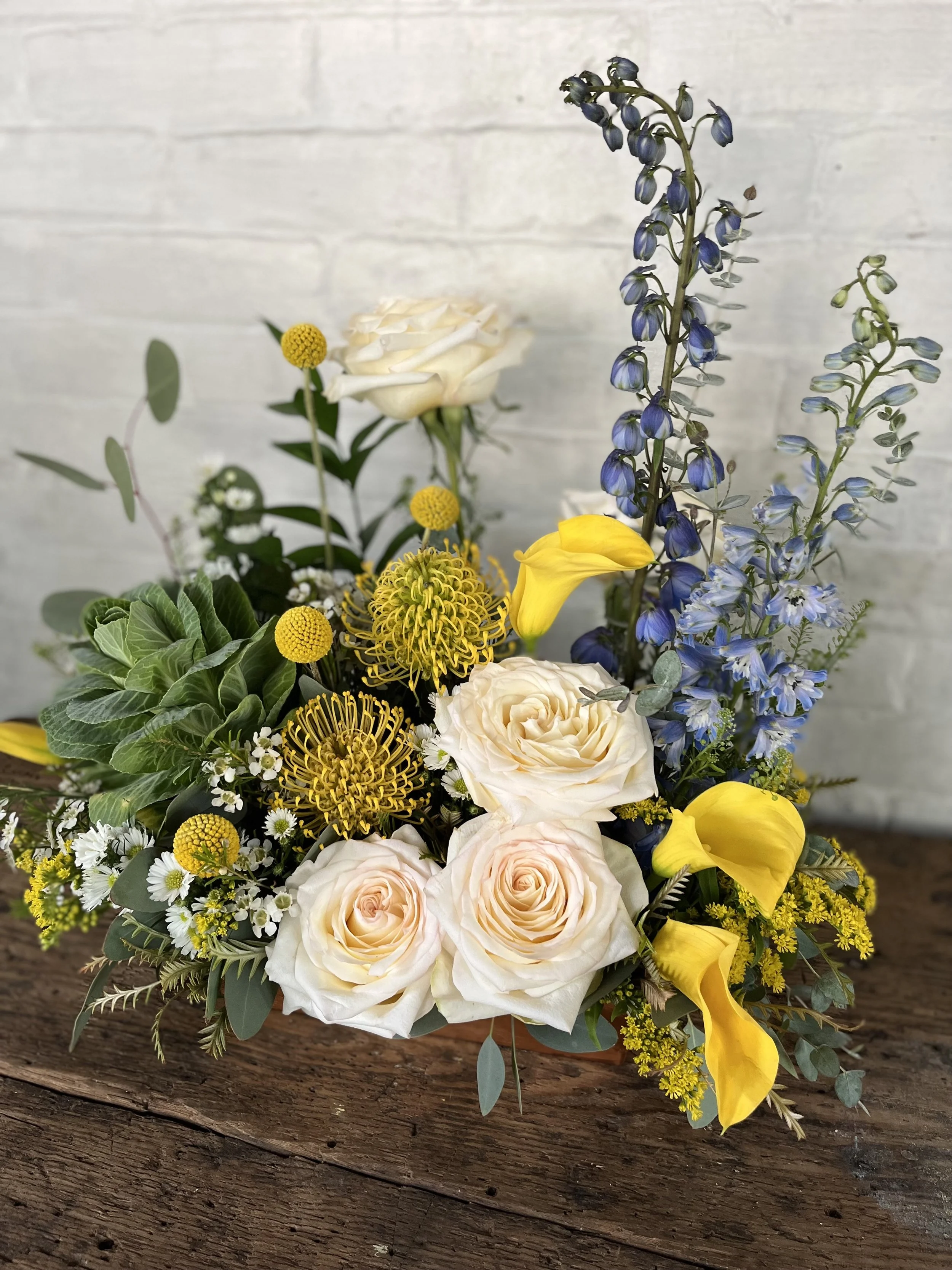 A floral arrangement consisting of white roses, yellow calla lilies, blue delphiniums, small white daisies, yellow billy balls, and green foliage, placed on a rustic wooden surface against a white brick wall background.