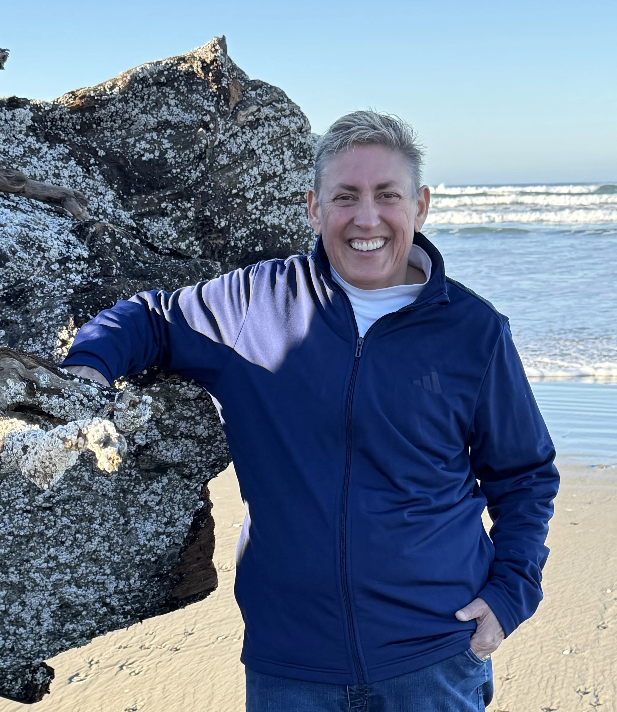 A smiling man standing on a beach next to a large, barnacle-covered driftwood log with ocean waves in the background.