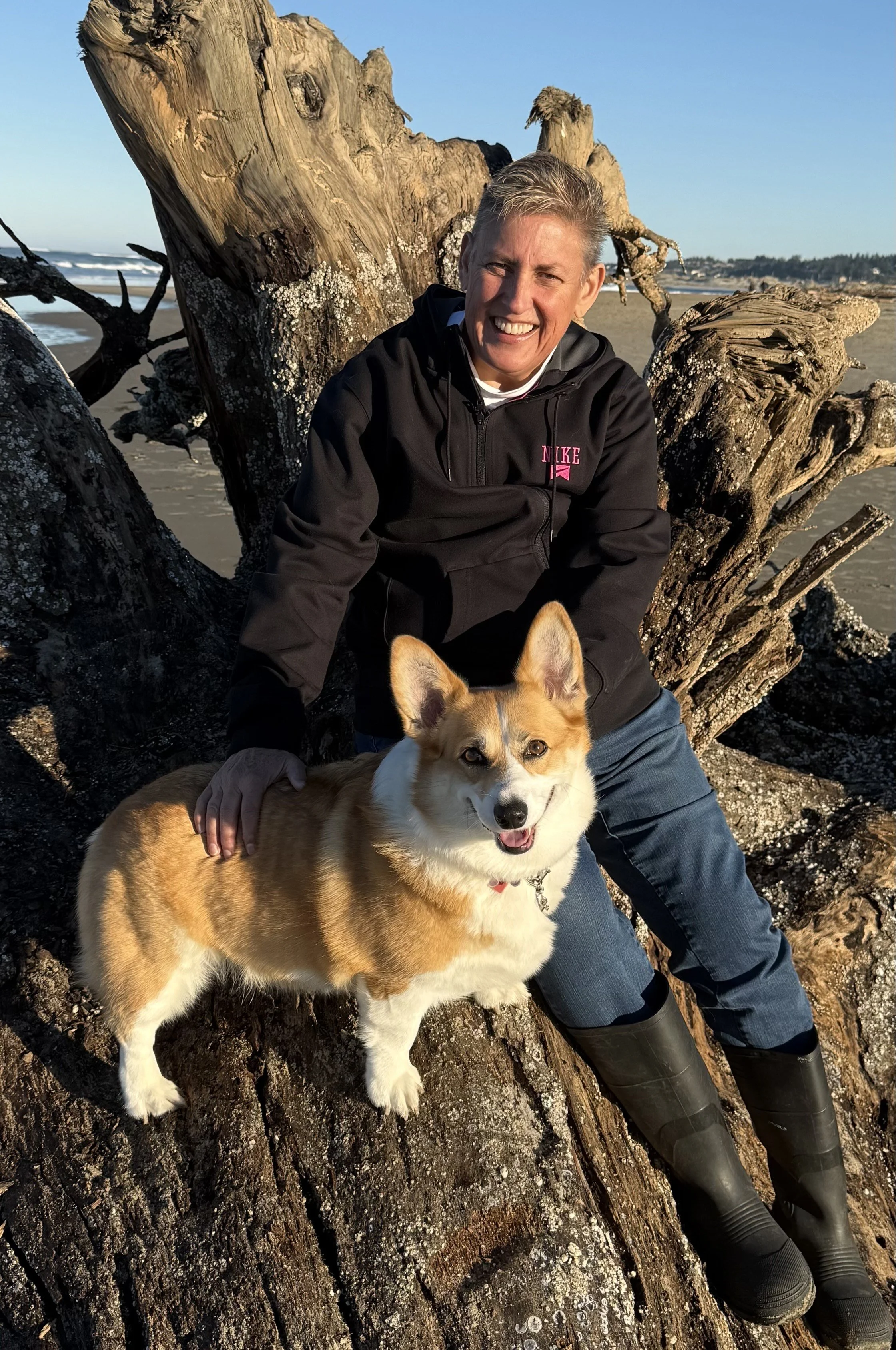 A woman with short gray hair smiling, wearing a black Nike jacket, sitting on a large piece of driftwood on the beach with a happy, tan and white corgi