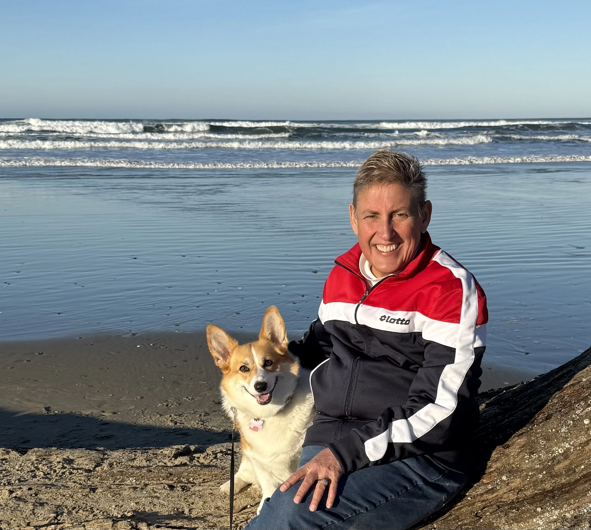 A man in a red, white, and black jacket sitting on a rock at the beach, smiling, with a corgi dog sitting beside him, ocean waves in the background, and clear sky.