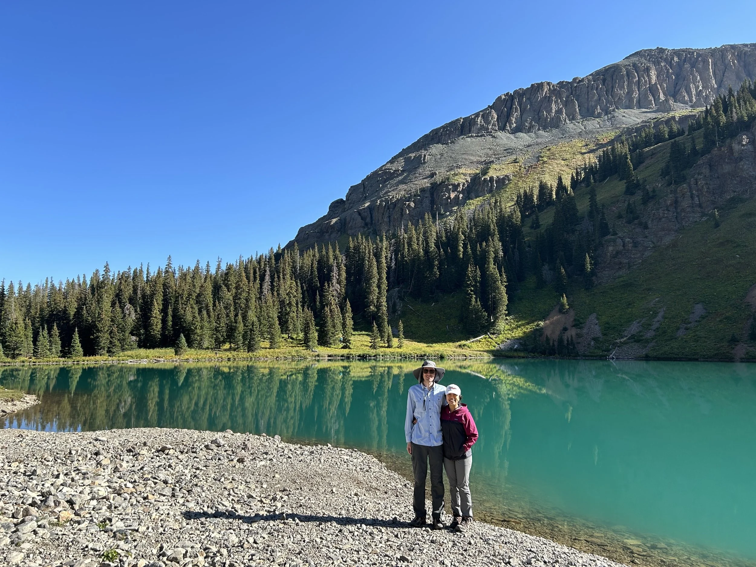 Veronica Longville & Jamie Longville standing by a turquoise lake surrounded by trees and mountains under a clear blue sky.