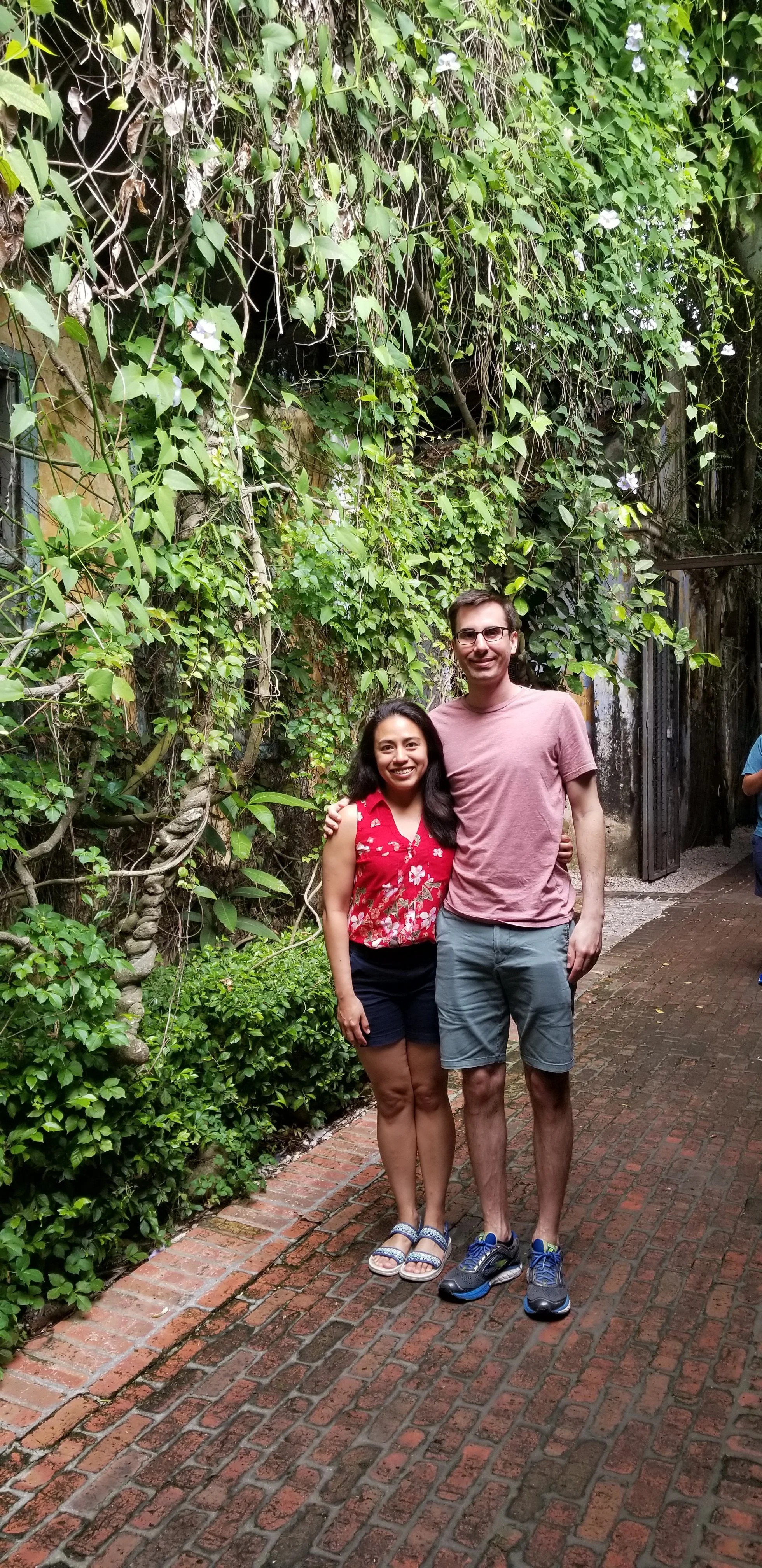 Veronica Longville & Jamie Longville posing together outdoors on a brick pathway, surrounded by lush green plants and climbing vines.